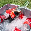 Botella de vidrio y latas de Coca‑Cola en una nevera portátil con hielo, sobre césped.