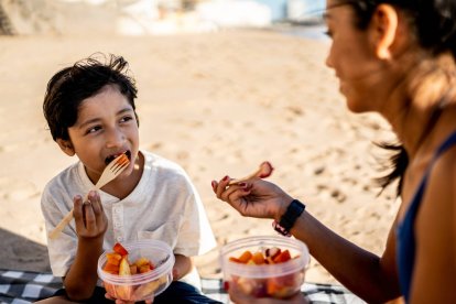 Niño comiendo macedonia pelada con su madre en la playa, expuestos al calor.