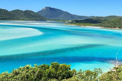 Vista panorámica de Whitehaven Beach en Australia, con aguas turquesa y arena blanca de sílice formando remolinos naturales entre colinas verdes.
