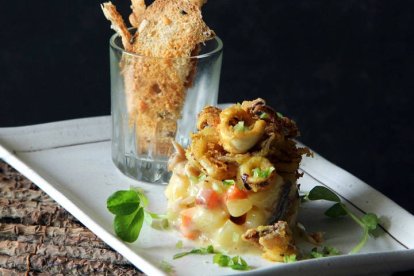 El chef presenta el plato en ración individual, coronado con chips de ajo y acompañado de tostas verticales, logrando un emplatado sofisticado y apetecible.