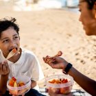 Niño comiendo macedonia pelada con su madre en la playa, expuestos al calor.