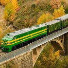 Locomotora verde del Expreso de Canfranc cruzando un puente ferroviario entre bosques otoñales del Pirineo aragonés.
