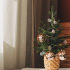 Un mini abeto natural decorado con elegancia sencilla: bolas doradas, luz suave y una cesta trenzada que eleva cualquier rincón navideño sin esfuerzo.