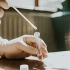 A hand wearing a latex glove holds a small clear tube and dips a cotton swab into the liquid. Space for copy.