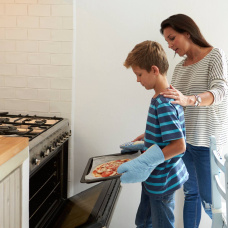 Una madre acompaña a su hijo mientras hornea una pizza en casa, en una escena cotidiana que refleja la preparación de una comida rápida y familiar.
