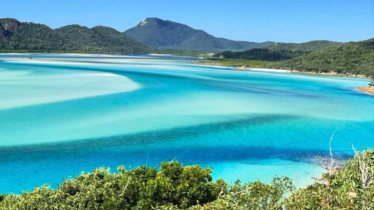 Vista panorámica de Whitehaven Beach en Australia, con aguas turquesa y arena blanca de sílice formando remolinos naturales entre colinas verdes.