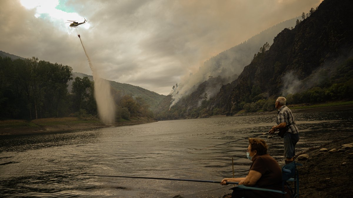 Un helicóptero recoge agua para ayudar con las labores de extinción del incendio mientras una pareja pesca, a 26 de agosto de 2025, en Aguas Mestas, Lugo.
